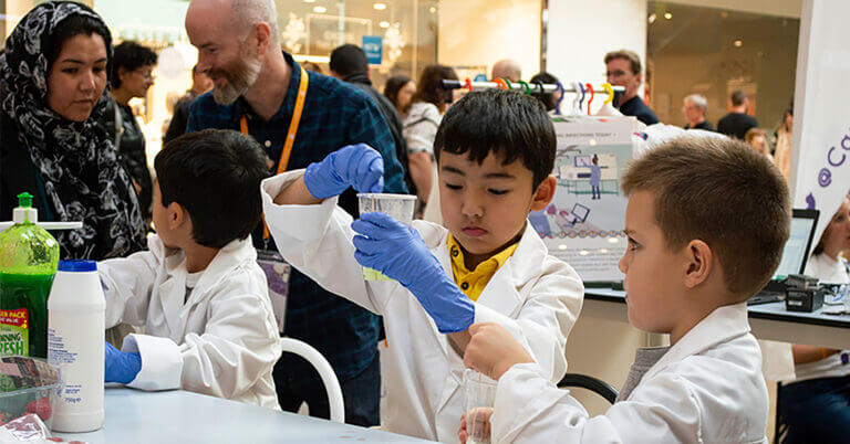 A group of young children wearing a lab coats and gloves as part of a hands-on public event.