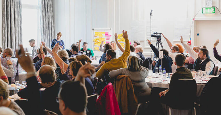 Photo of a previous Citizens' Jury run by Involve. A room full of participants taking part in a discussion, some with their hands up. There is a board covered in yellow and pink post-it notes where notes have been written by jury memebers.