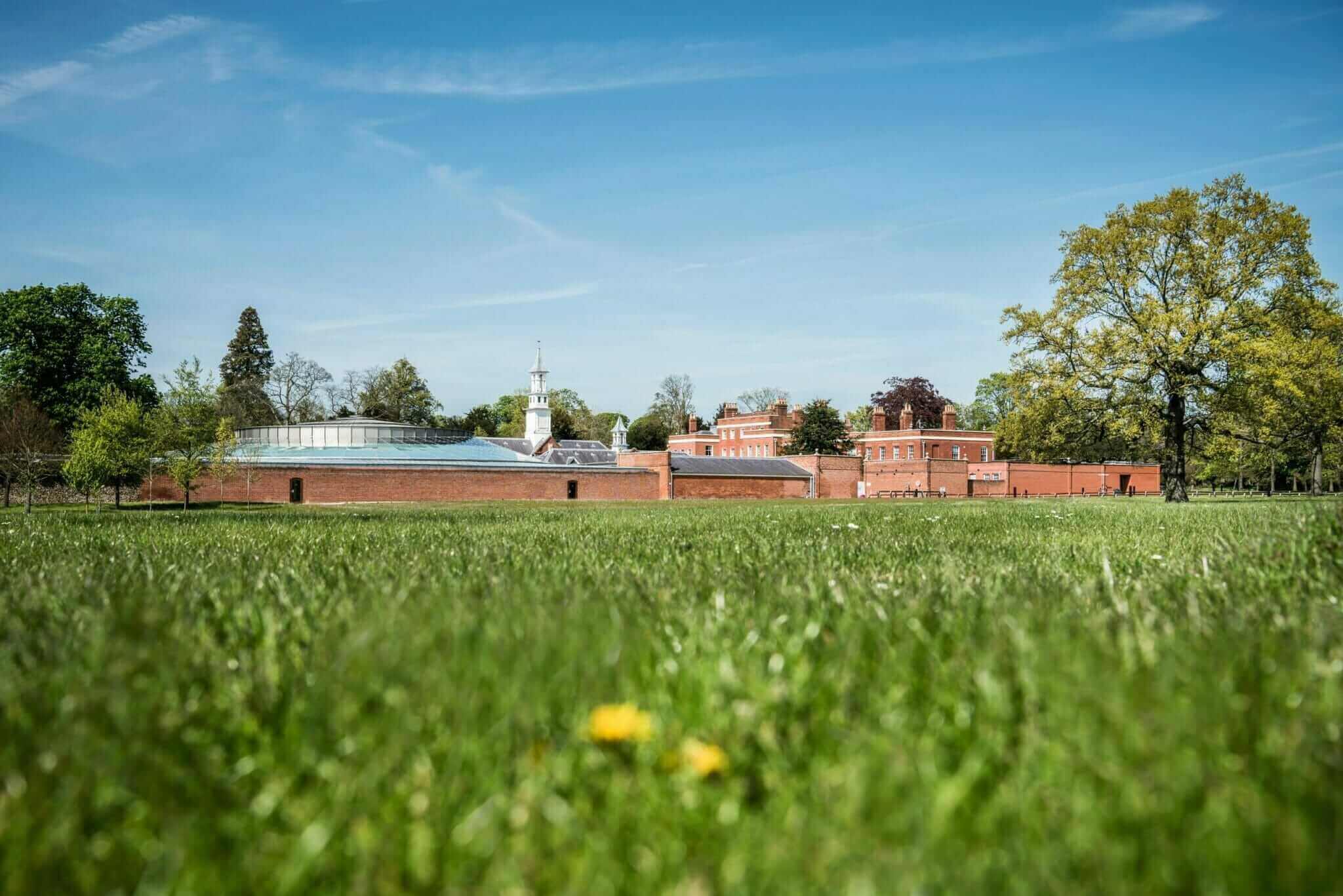 A colour photo of green grass on the Wellcome Genome Campus, with the Conference Centre red brick buildings in the background.