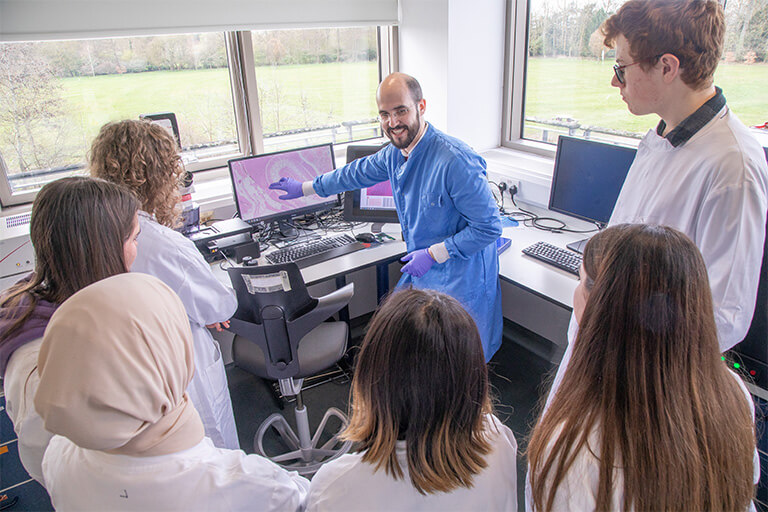 Group of sixth from students wearing white lab coats watching smiling scientist explain what's on the computer screen