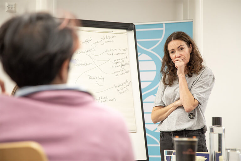 Woman looking thoughtful by a flip chart listening to person speaking (we see the back of their head only)