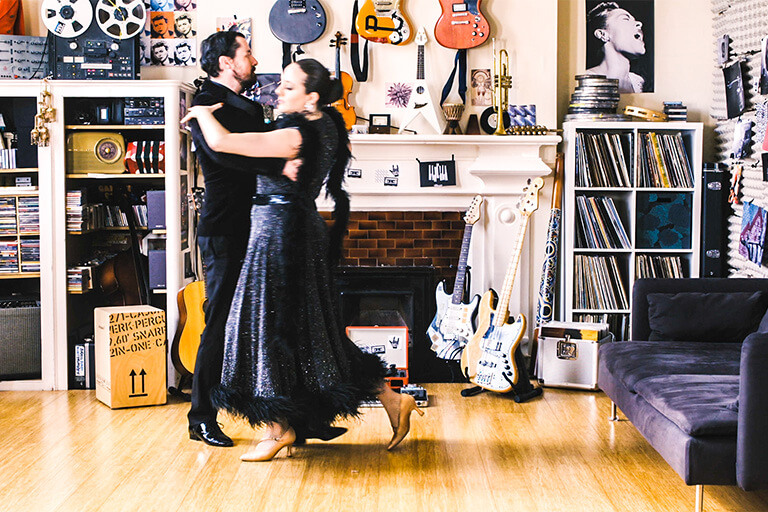 A couple dressed in black dancing a formal dance, in a living room setting with electric guitars in the background. Still image from 'What is a gene?' film in the Music of Life series.