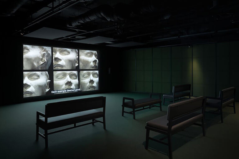 Photo of an exhibition room at the Wellcome Collection with a screen comprising of 6 screens and three benches for viewers.