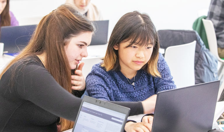 Two young women are talking and looking at a laptop in a workplace.