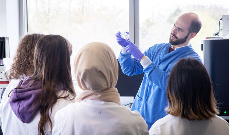 A male scientist is showing a group of young female students a sample in a beaker.