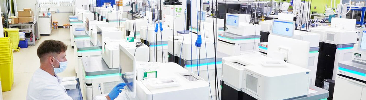 A photo of a man working in a DNA sequencing lab, full of sequencing machines. He is wearing a mask.