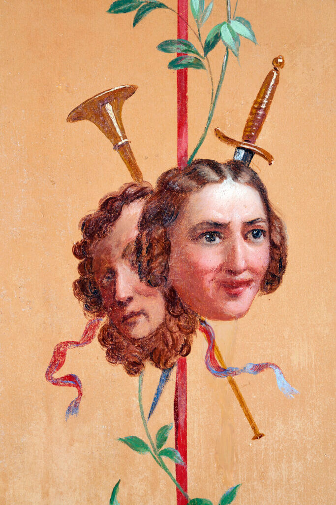 Detail in a mural in the Pompeiian Room in Hinxton Hall showing the heads of Julia Flower and Edward Humphrys Greene de Freville. Peach coloured background, and a crossed sword and herald's trumpet are behind the heads.