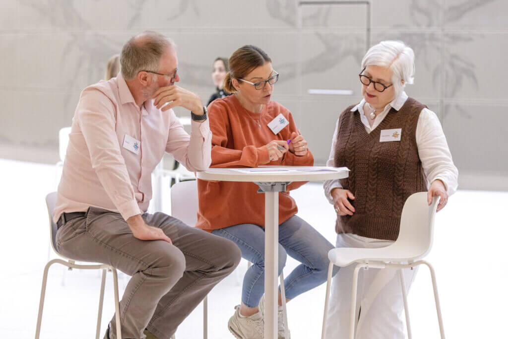 Two women and a man are sat around a small table in deep discussion, looking at a leaflet on the table-top.