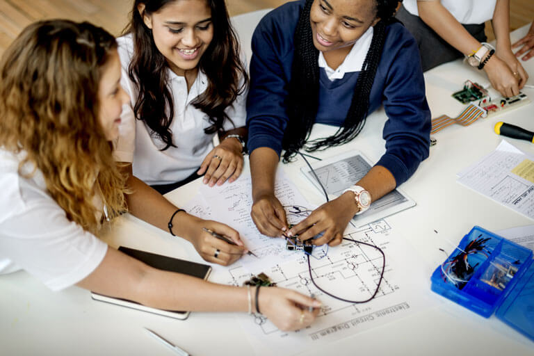 Three teenage girls are sat working and smiling, they are wearing school uniform.